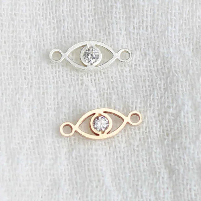 Two gold eye-shaped jewelry pieces with embedded stones on a light gray background